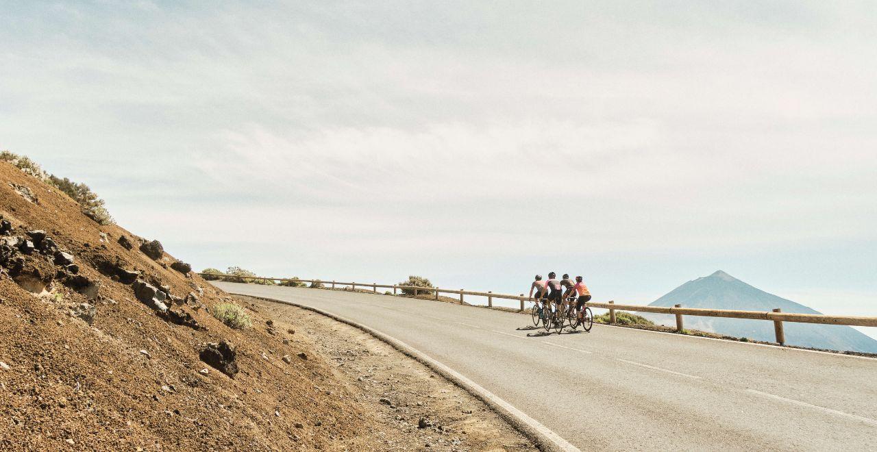 Group of cyclists riding uphill on a paved road in Tenerife, with Mount Teide visible in the background and a clear sky.