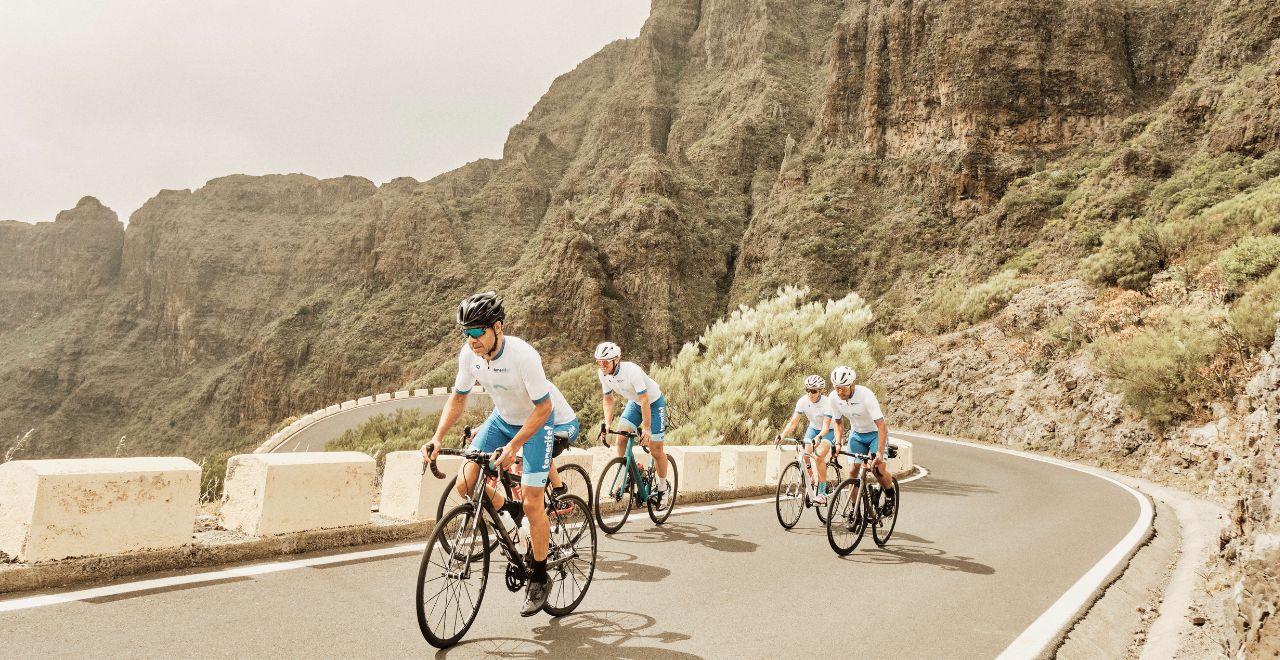 Cyclists riding on a scenic road in Masca, Tenerife, surrounded by towering cliffs and lush vegetation.