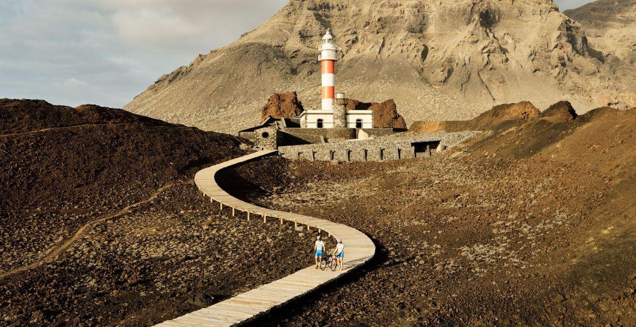 Cyclists walking towards Punta de Teno lighthouse on a winding path through volcanic terrain in Tenerife.