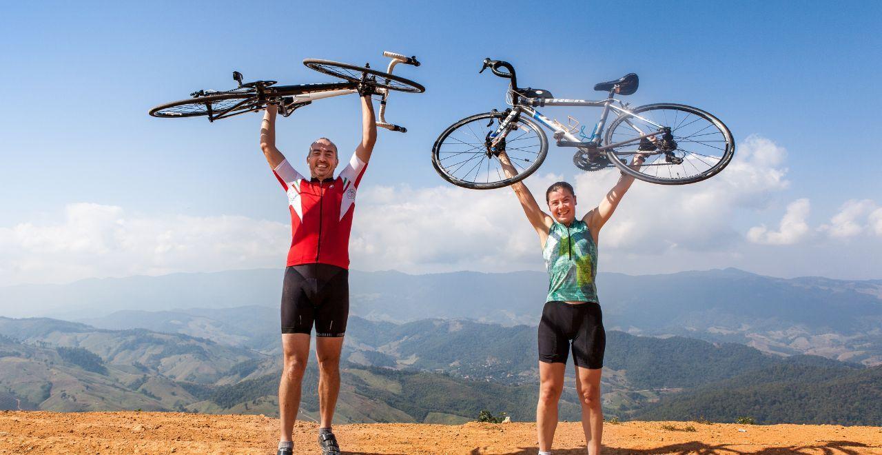 Two cyclists celebrating on a mountain top, holding bikes overhead
