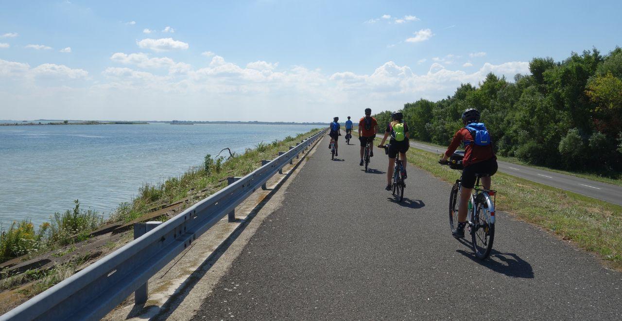 Group of cyclists riding along a paved road by a large body of water under a blue sky with clouds