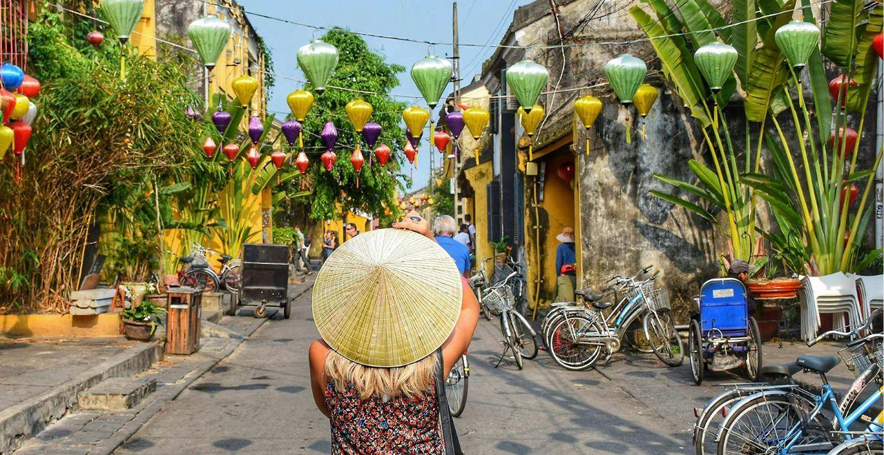 Woman in a Vietnamese conical hat photographing colorful lanterns in Hoi An street.