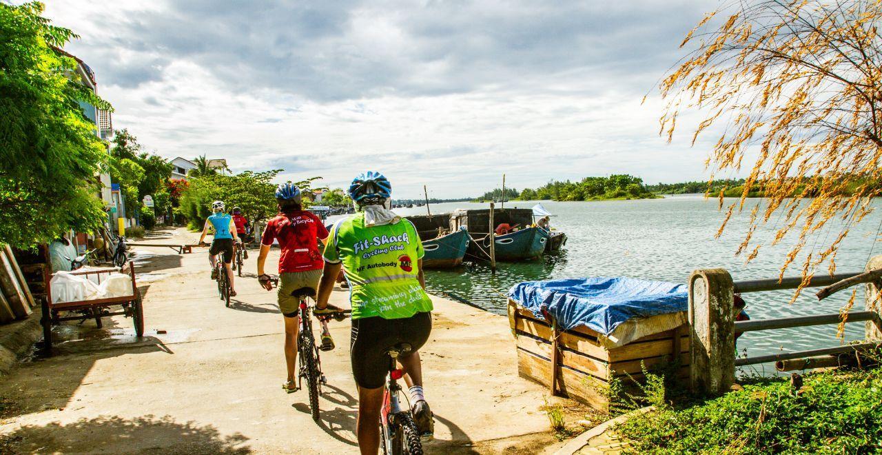 Cyclists riding beside a serene river in Vietnam, interacting with local life and watercraft on their journey.