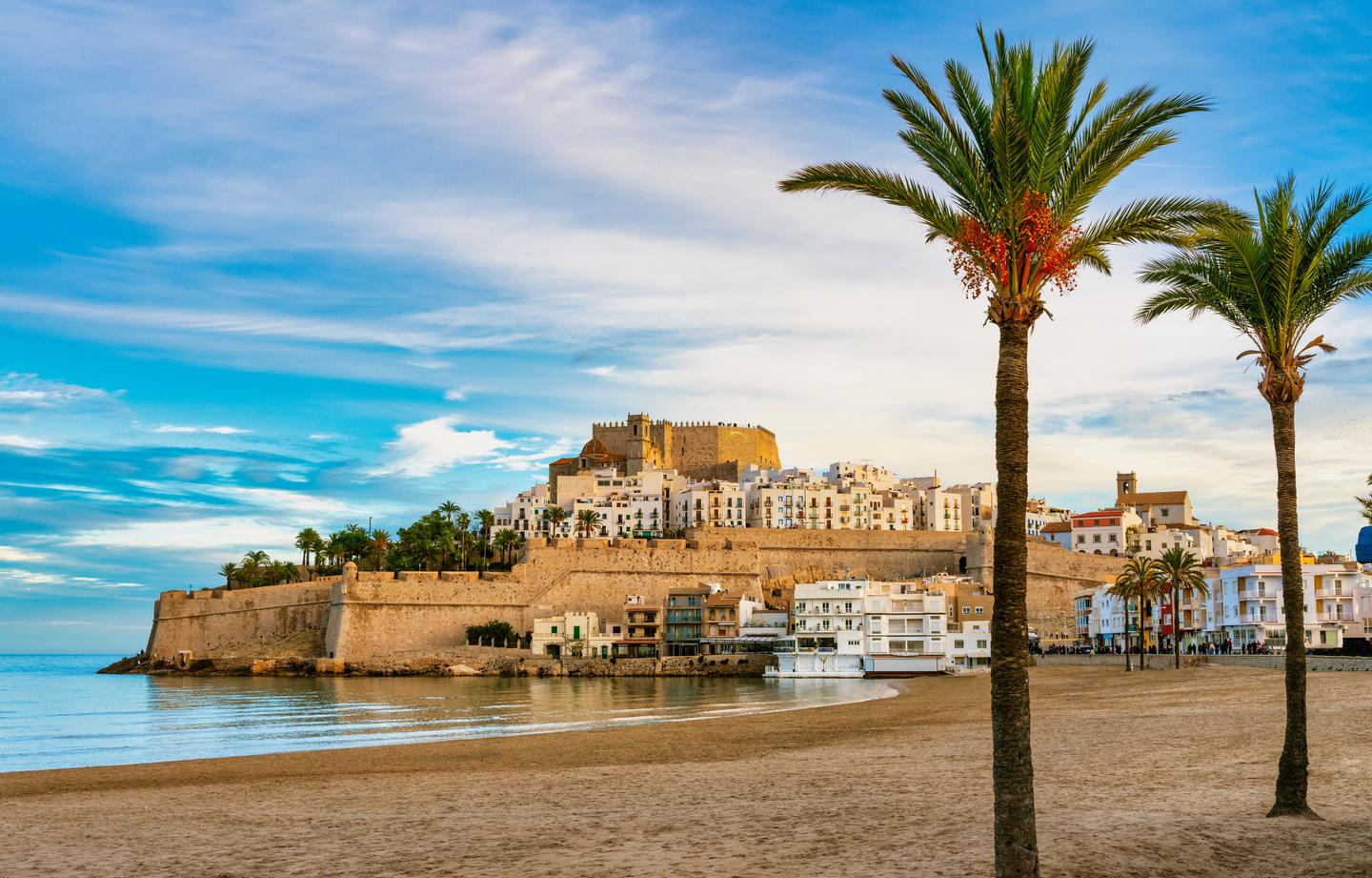 The historic fortress of Peñíscola in Castellón, Spain, standing majestically on a rocky headland. The sandy beach and palm trees in the foreground contrast with the ancient walls and whitewashed buildings, under a blue sky with scattered clouds.