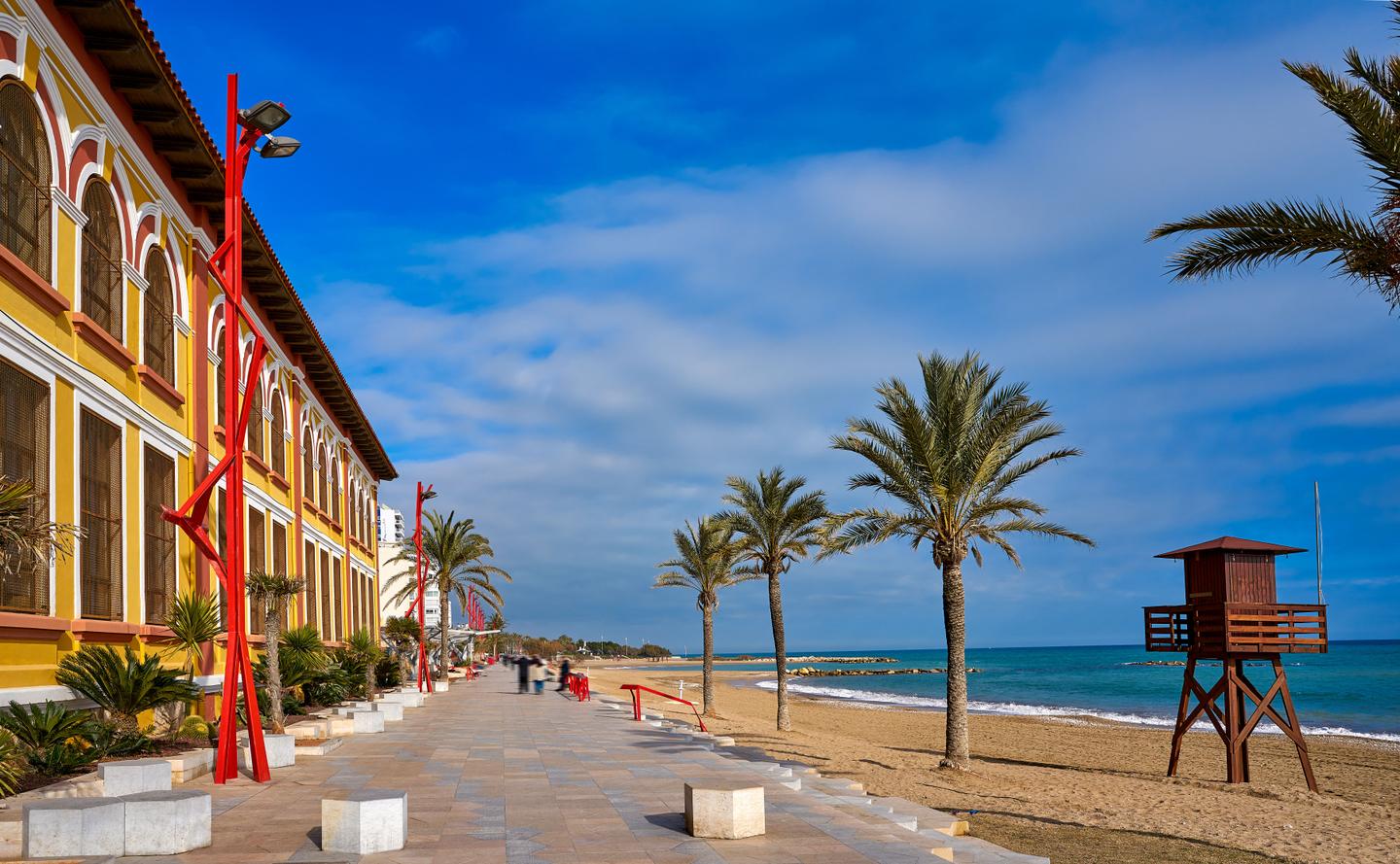 A vibrant seaside promenade in Castellón, Spain, featuring colorful buildings with arched windows, red lamp posts, and palm trees lining the walkway. The sandy beach and calm sea are visible, creating a picturesque coastal scene.