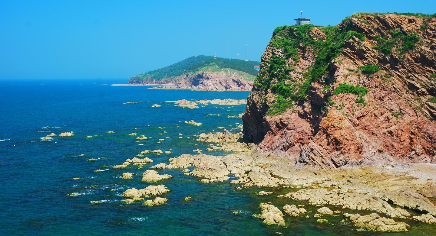 Scenic view of a rocky cliff by the sea with clear blue water and a bright blue sky. The coastline features scattered rocks and lush green vegetation atop the cliffs, creating a picturesque coastal landscape.