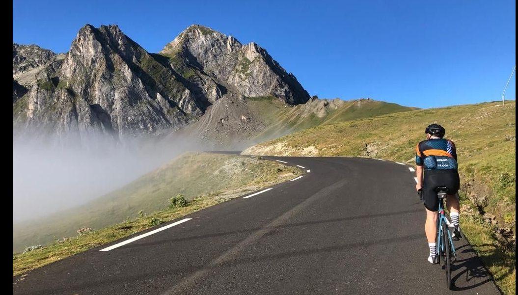 A cyclist ascends the Col du Tourmalet on a clear day, with rugged mountain peaks in the background and a layer of fog settled in the valley below.