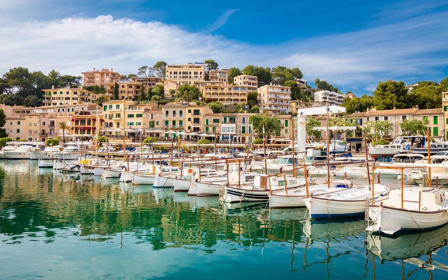 Scenic view of Port de Sóller marina in Mallorca, with boats docked in the clear turquoise water and colorful hillside buildings under a bright blue sky