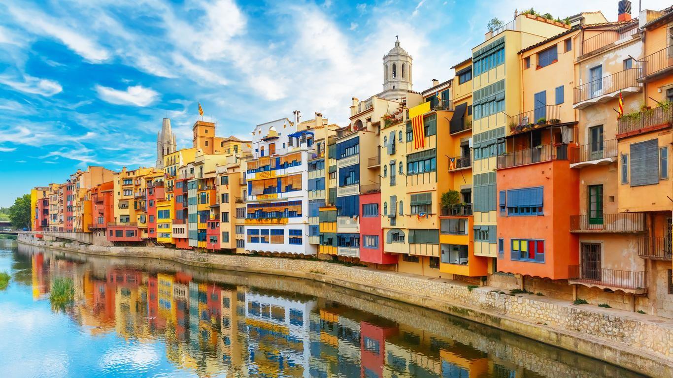 Colorful buildings along the Onyar River in Girona, with a clear blue sky and reflections on the water.