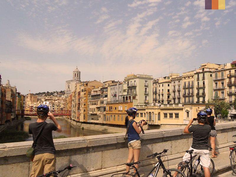 Group of cyclists taking photos on a bridge in Girona, with colorful buildings and a river in the background under a clear sky.