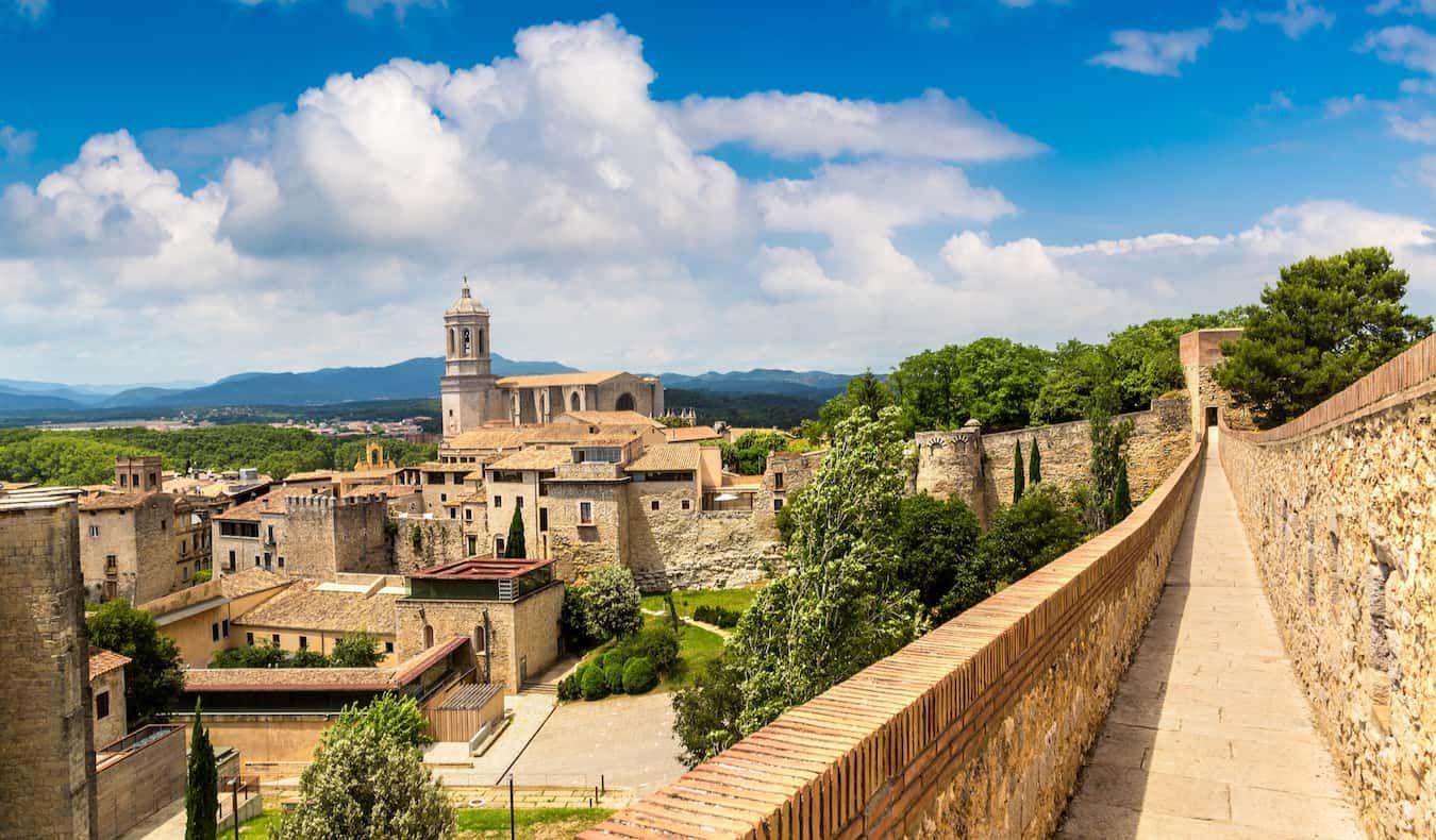 View of Girona's historic old town, featuring the Girona Cathedral and ancient stone walls under a bright blue sky with scattered clouds.