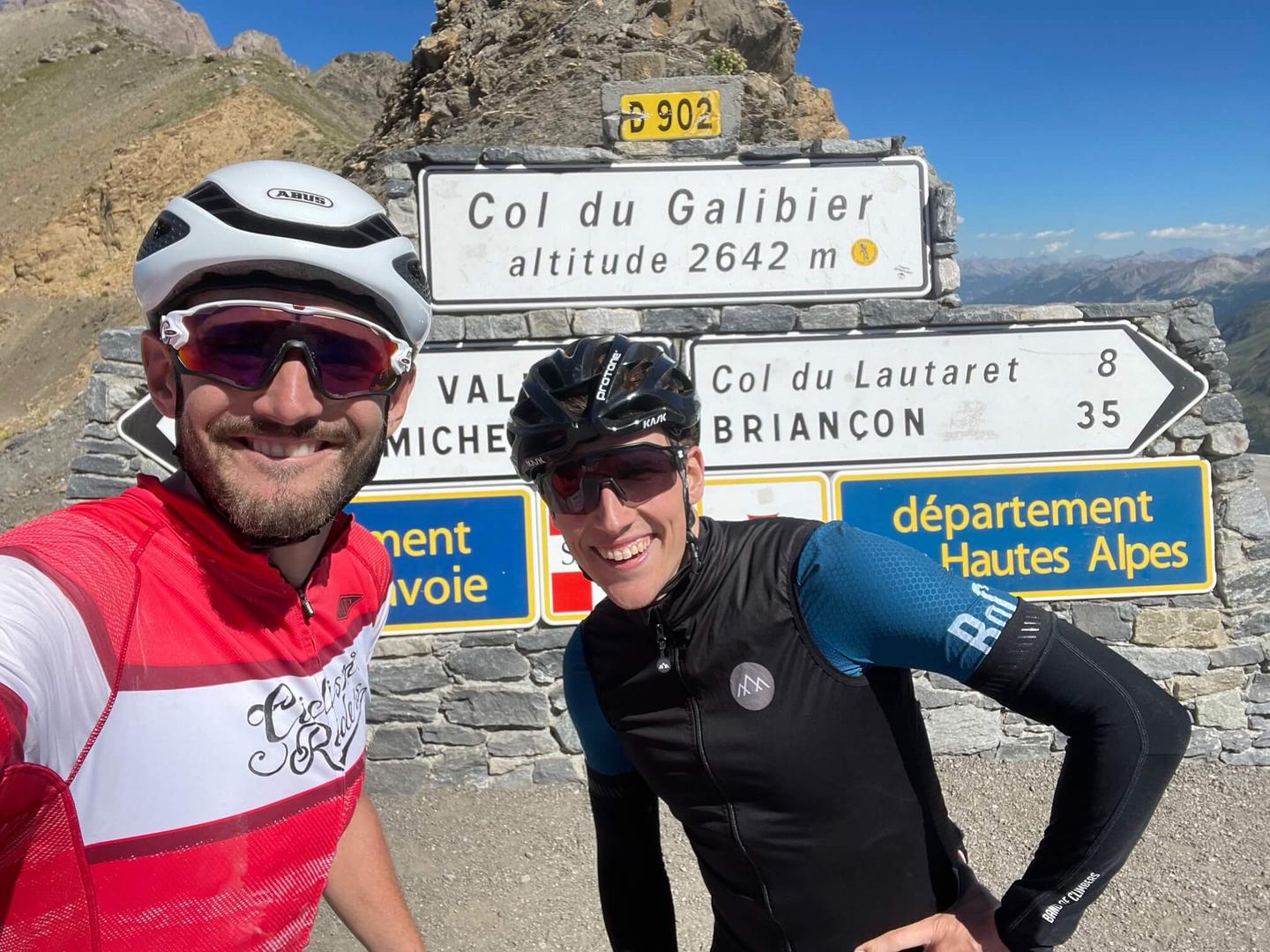 Two cyclists celebrating at Col du Galibier altitude sign, Hautes Alpes, France, scenic mountain background.