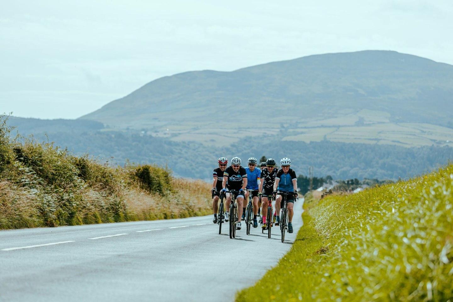 A group of cyclists riding on a road through the countryside in the Isle of Man, with green fields and hills in the background.