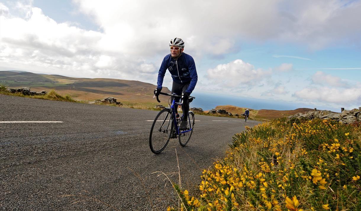 A solo cyclist climbs a road on the Isle of Man, surrounded by rolling hills and wildflowers. The sea is visible in the background under a partly cloudy sky.
