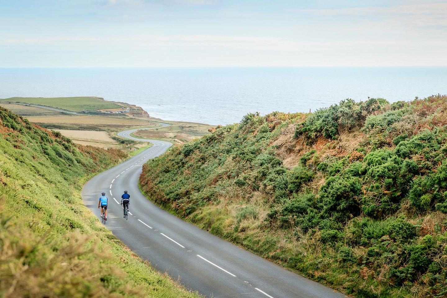 Two cyclists riding on a winding coastal road in the Isle of Man, surrounded by lush green hills with a view of the ocean in the background.