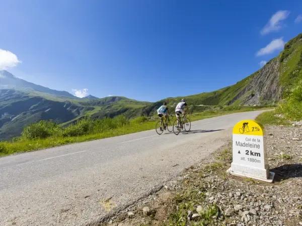 Two cyclists riding up a steep road in Col de la Madeleine, with a milestone indicating 2 km to the summit at an altitude of 1880m, surrounded by lush green mountains under a clear blue sky.