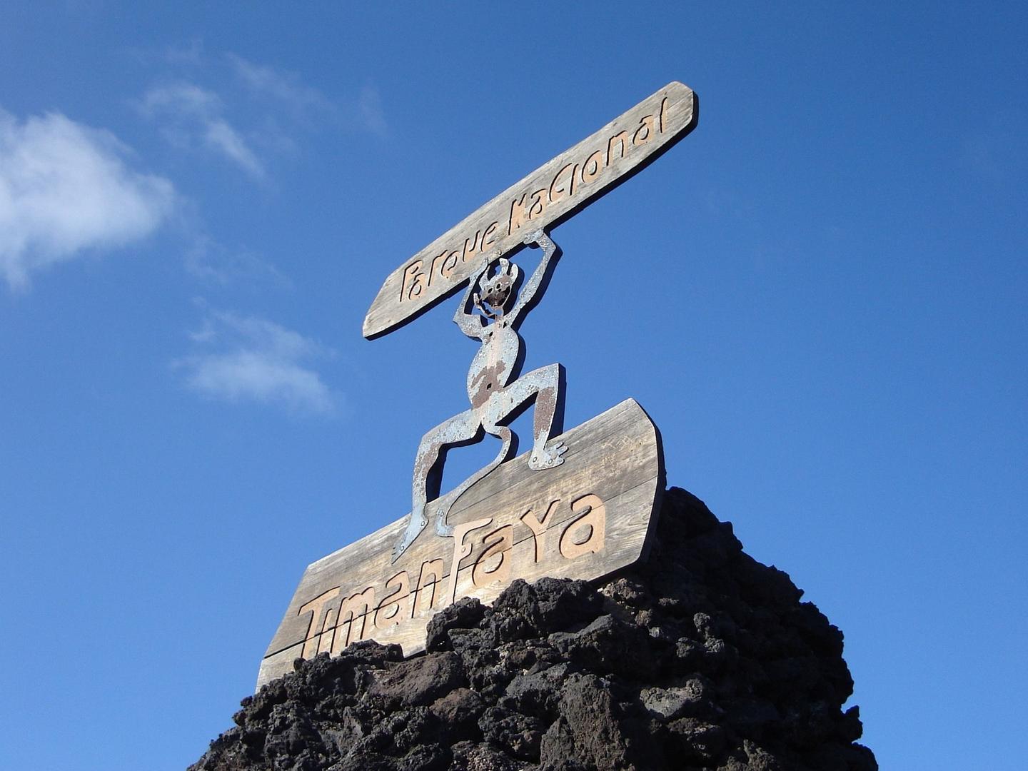The iconic entrance sign of Timanfaya National Park in Lanzarote, featuring a stylized figure holding a board with the park's name, set against a clear blue sky.