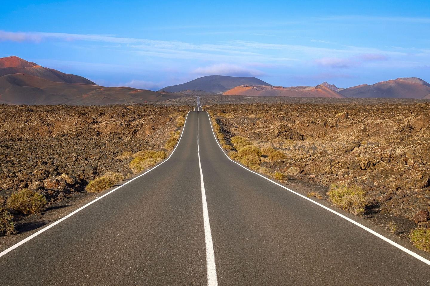 A long, straight road cutting through the volcanic landscape of Lanzarote, with rocky terrain and distant mountains under a clear blue sky.