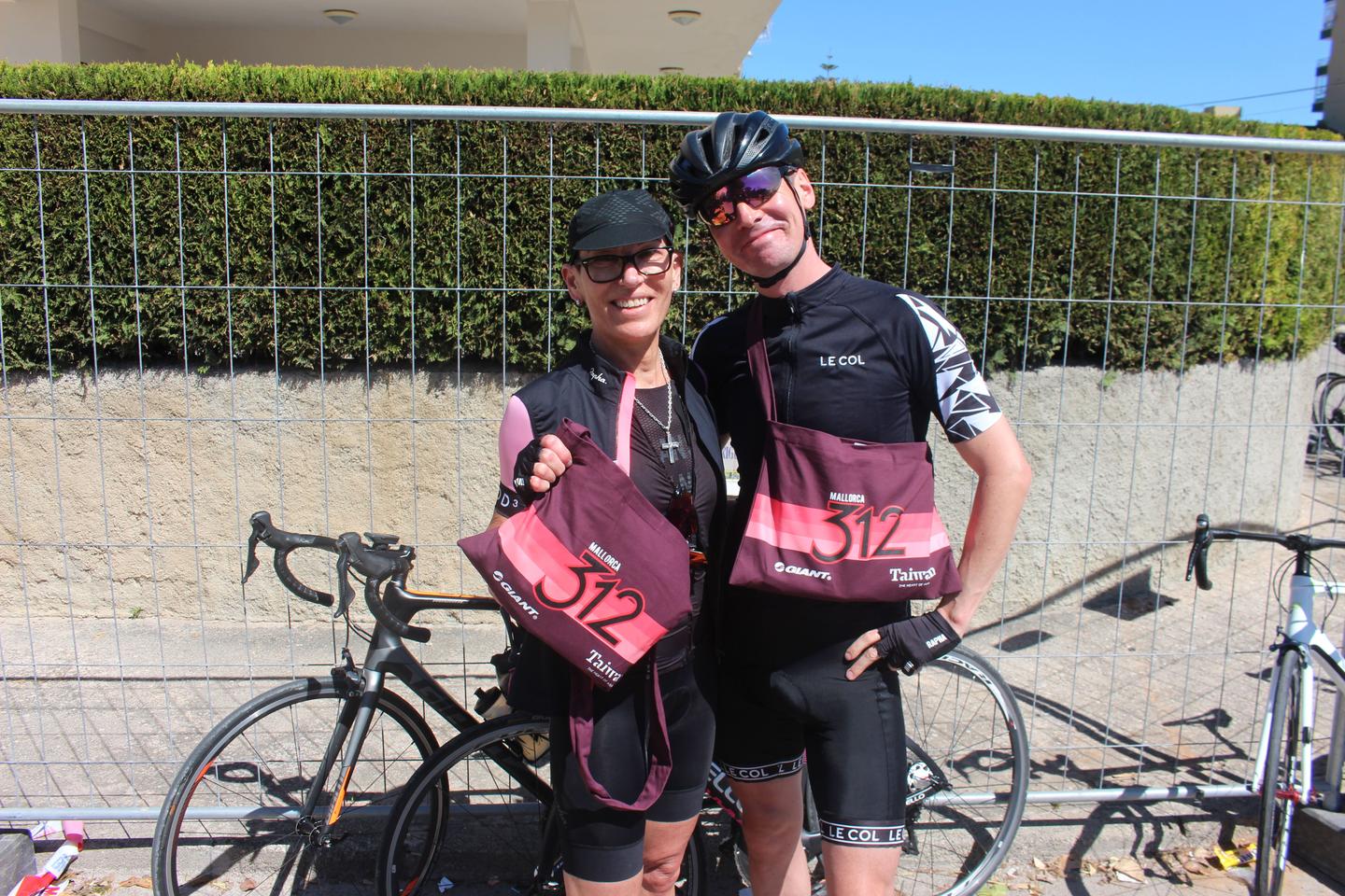 Two cyclists smiling and holding race bags at the Mallorca 312 event, standing in front of a fence with their bicycles nearby