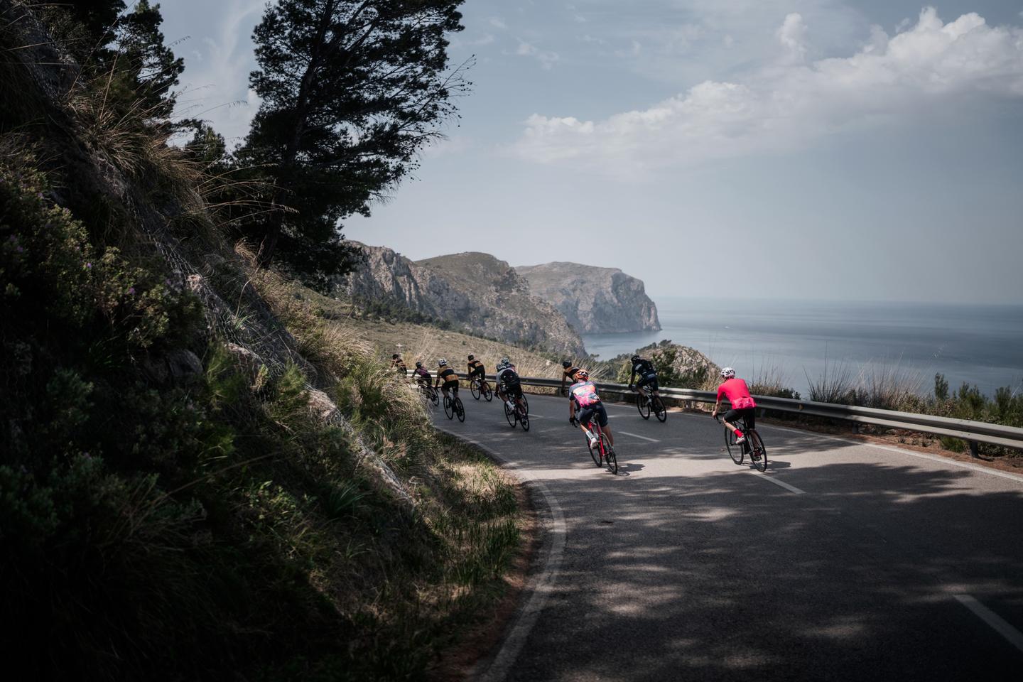 Group of cyclists riding on a coastal mountain road, with a view of cliffs and the ocean in the background. The road is surrounded by lush vegetation and trees under a cloudy sky.
