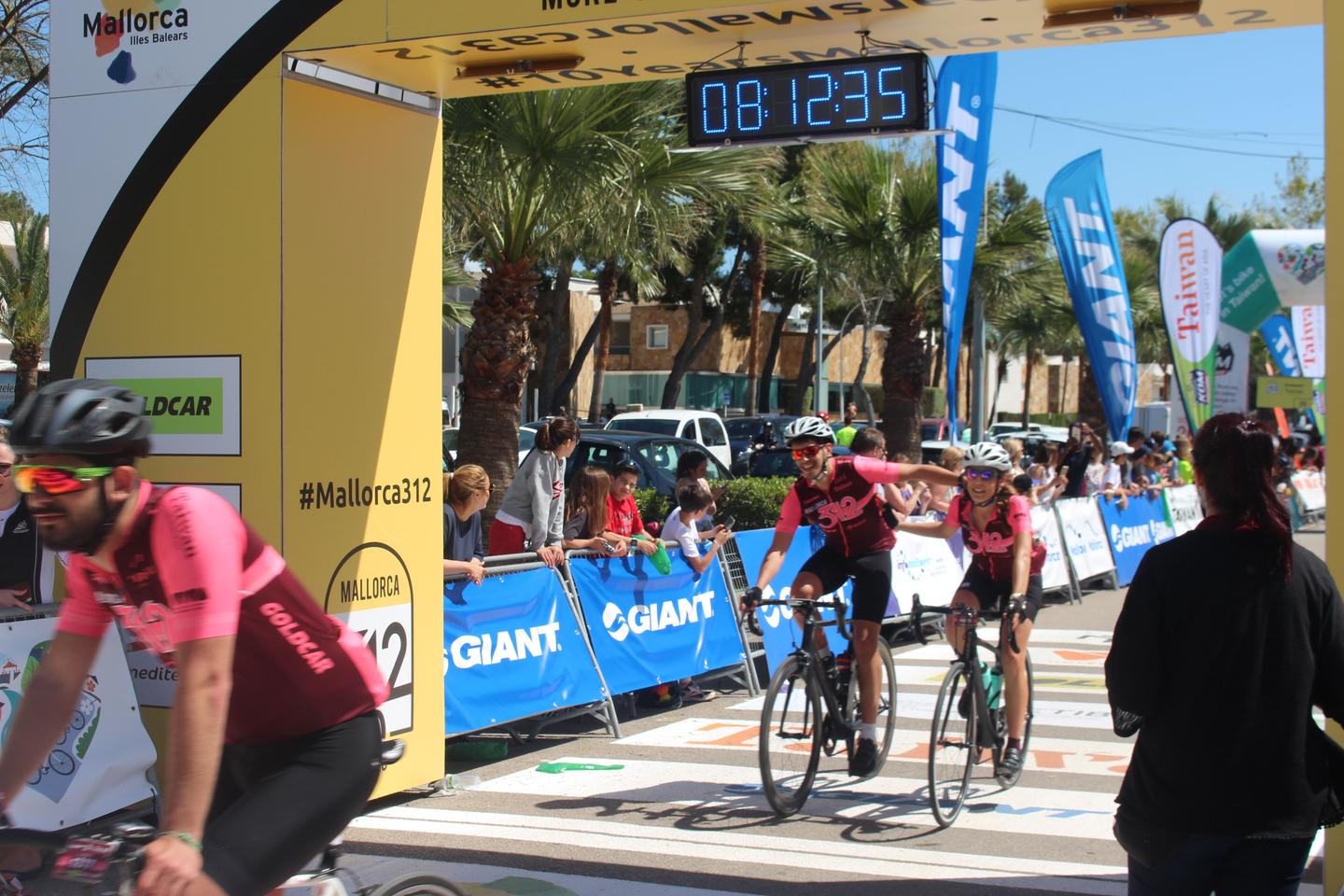 Cyclists crossing the finish line of the Mallorca 312 race, with spectators and a large digital timer overhead.