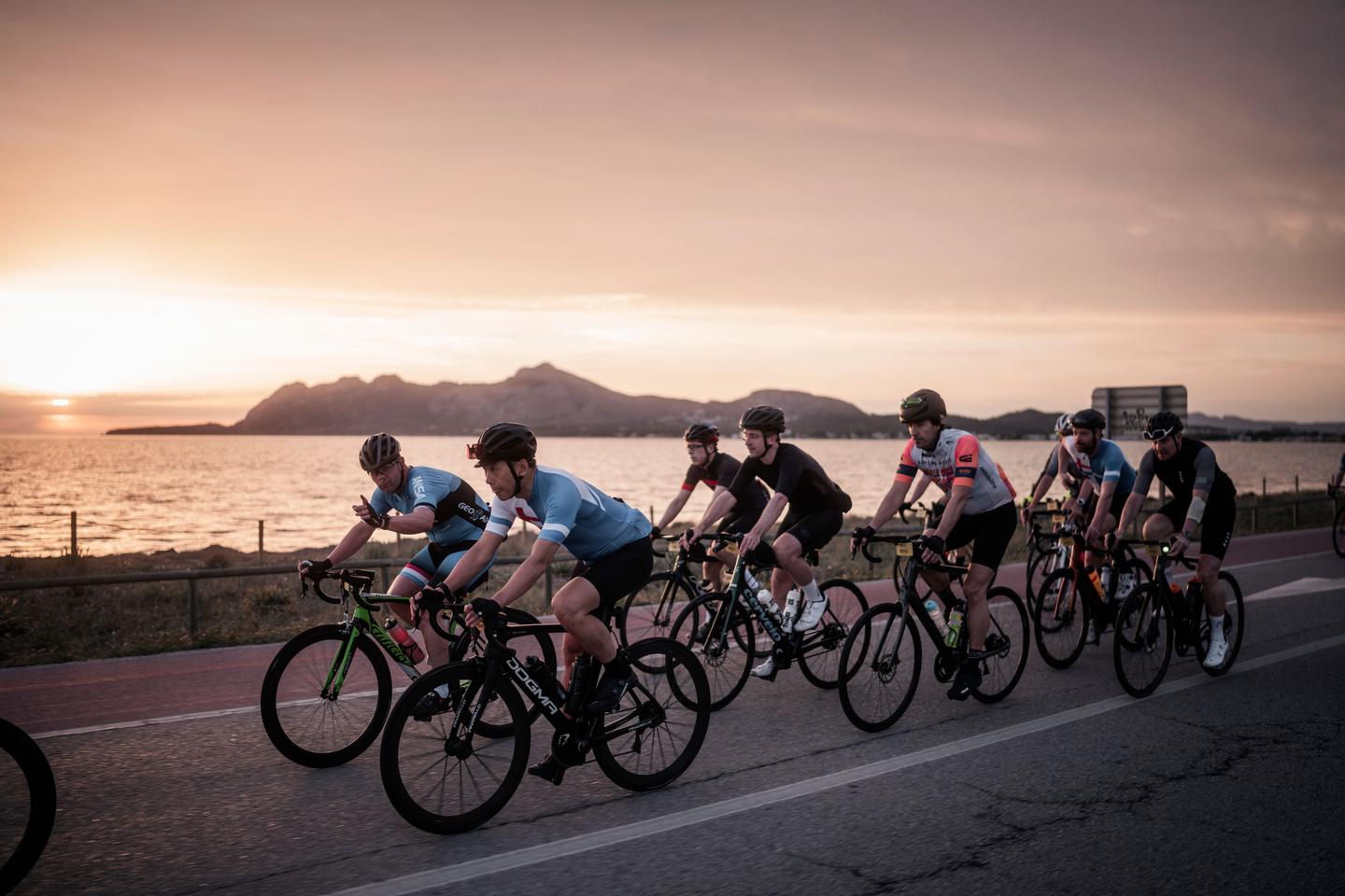 Group of cyclists riding along a coastal road at sunset, with mountains and the sea in the background. The sky is painted with warm colors, creating a serene and picturesque scene.