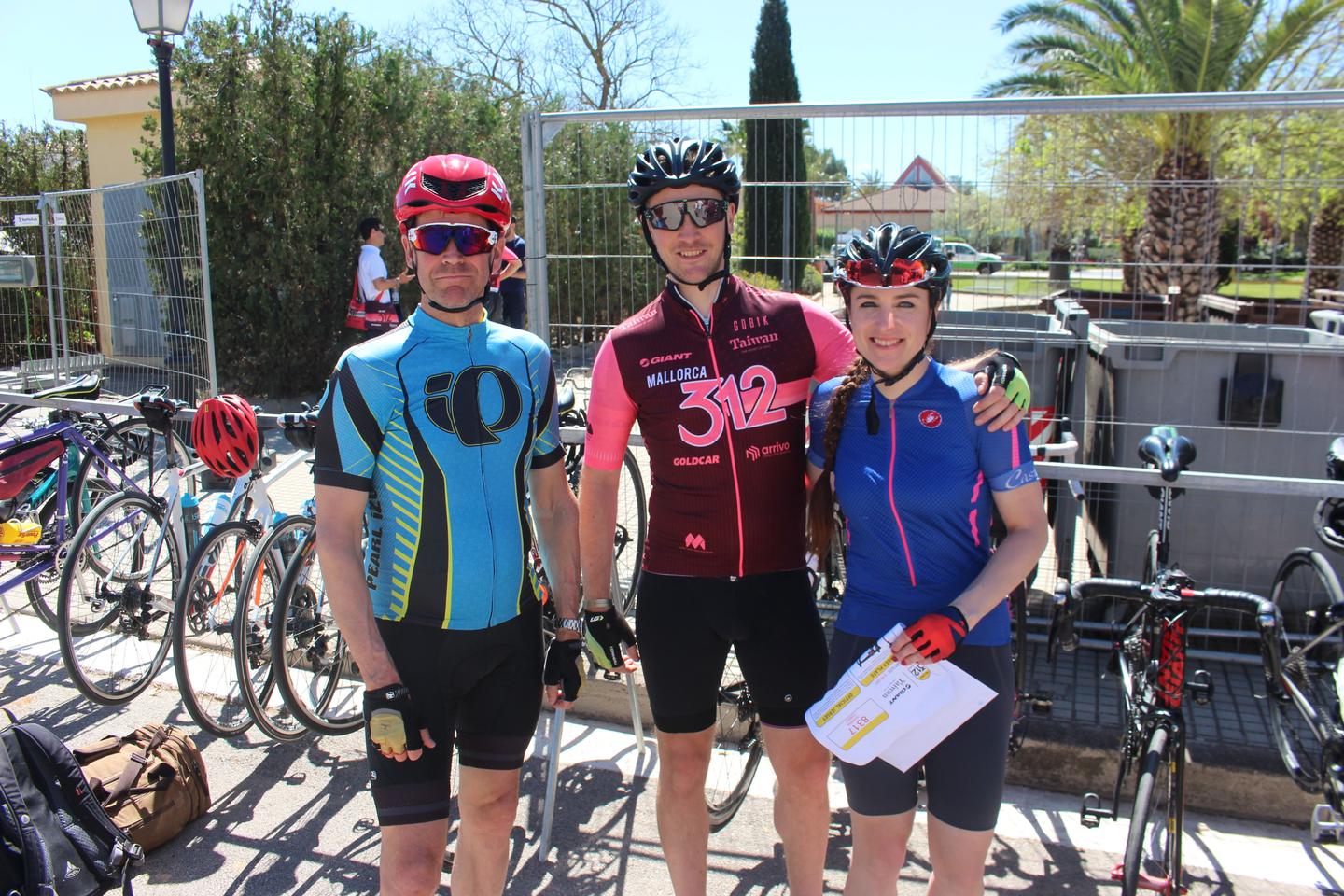 Three cyclists posing for a photo at the Mallorca 312 event, wearing colorful jerseys and helmets, with bicycles in the background.
