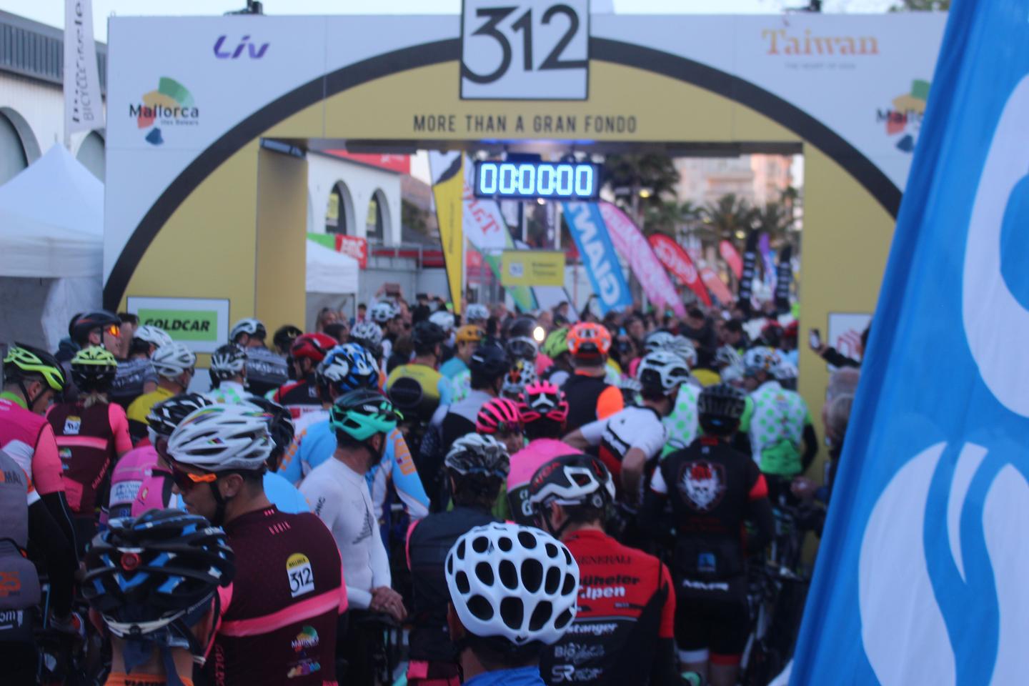 Cyclists gathered at the starting line of the Mallorca 312 race, with a large banner and digital timer displaying 00:00:00.