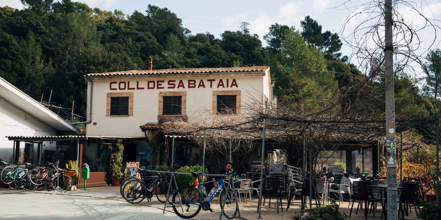 Cyclists and bikes parked outside a rustic building with a sign reading 'Coll de Sa Bataia,' surrounded by greenery.
