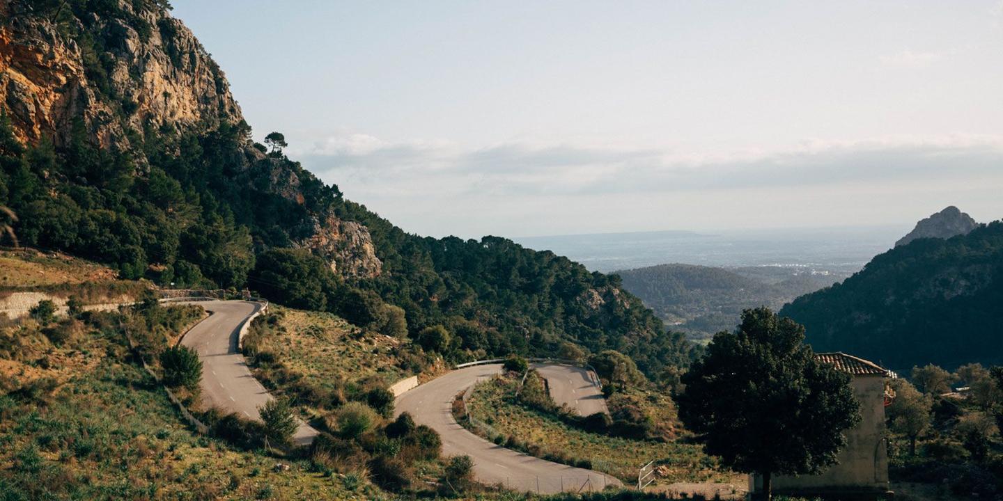 Winding mountain road surrounded by green hills and rocky cliffs in Mallorca under a clear sky.