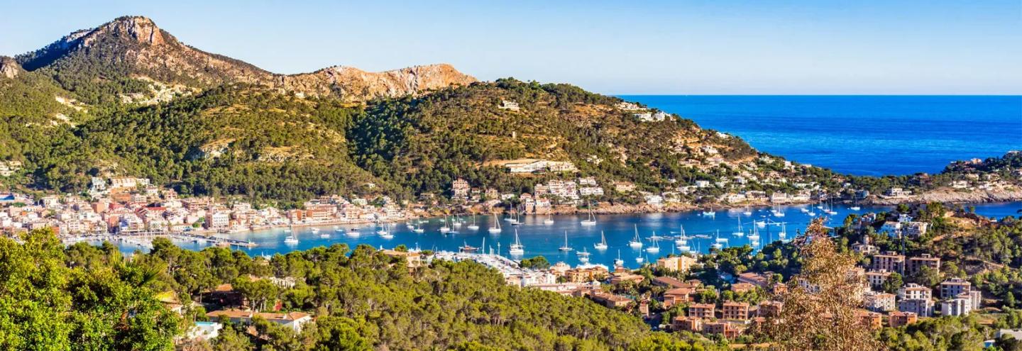 A panoramic view of a coastal town in Mallorca with hills, forests, and a harbor filled with boats, showcasing the scenic beauty of the island's cycling routes.