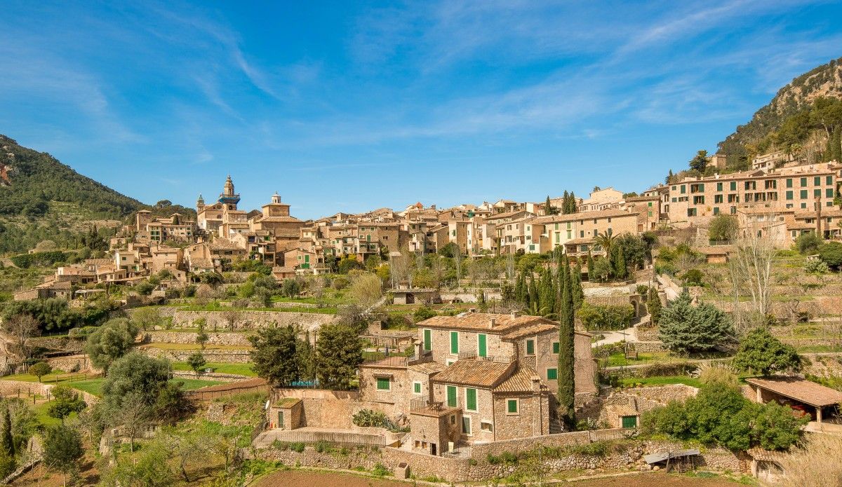 View of the village of Valldemossa nestled in the Tramuntana mountains, featuring terraced gardens and traditional stone houses under a clear blue sky.