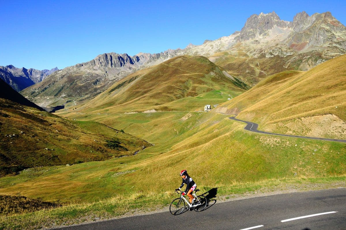 A cyclist climbing the Col du Glandon, surrounded by expansive green hills and rugged mountains under a clear blue sky.