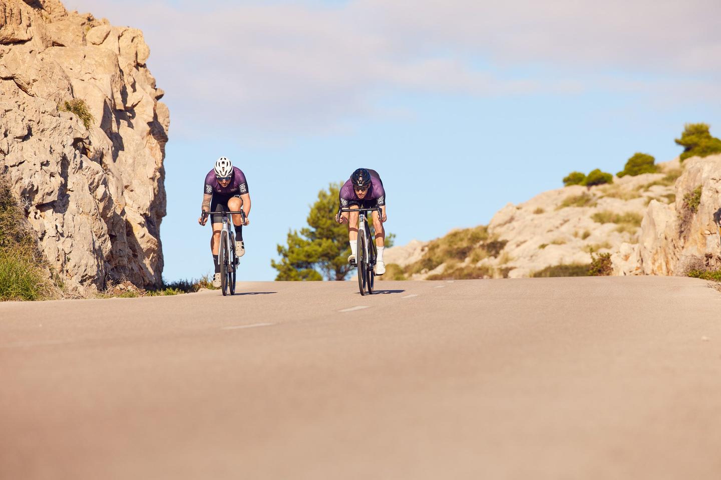 Two cyclists ride uphill on a sunny day, flanked by rocky outcrops and sparse vegetation, with a clear blue sky in the background.