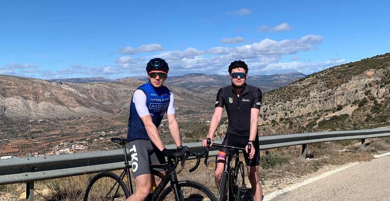 Two cyclists posing with their bikes on a mountain road with a panoramic view of the landscape.