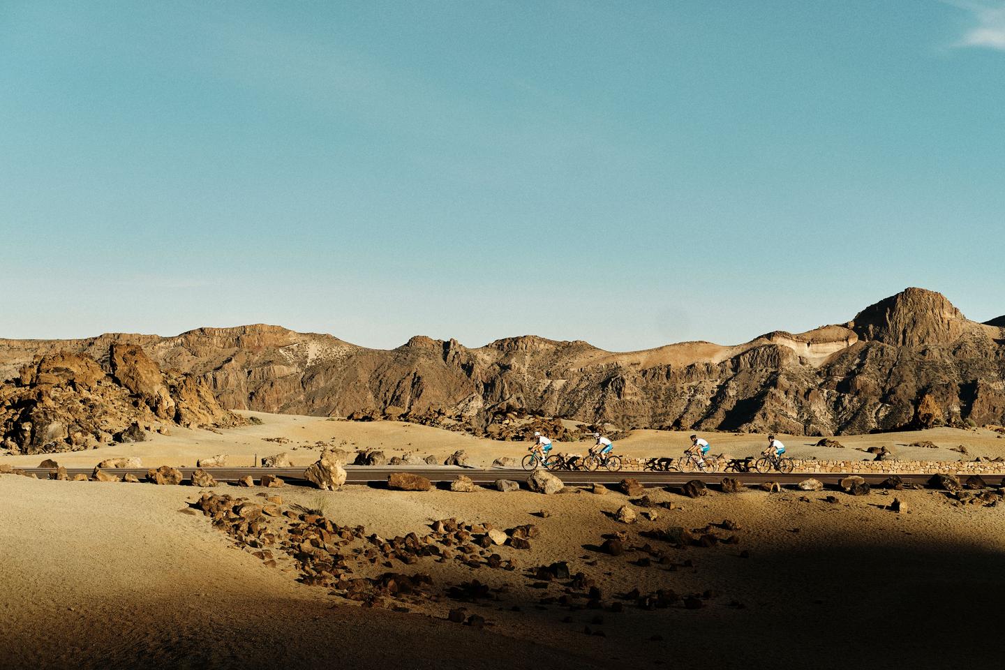 Group of cyclists riding through a rocky desert landscape with rugged mountains in the background under a clear blue sky