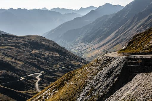 Winding roads of the Col du Tourmalet in the French Pyrenees snake through rugged mountains and lush valleys under a hazy, mountainous backdrop.