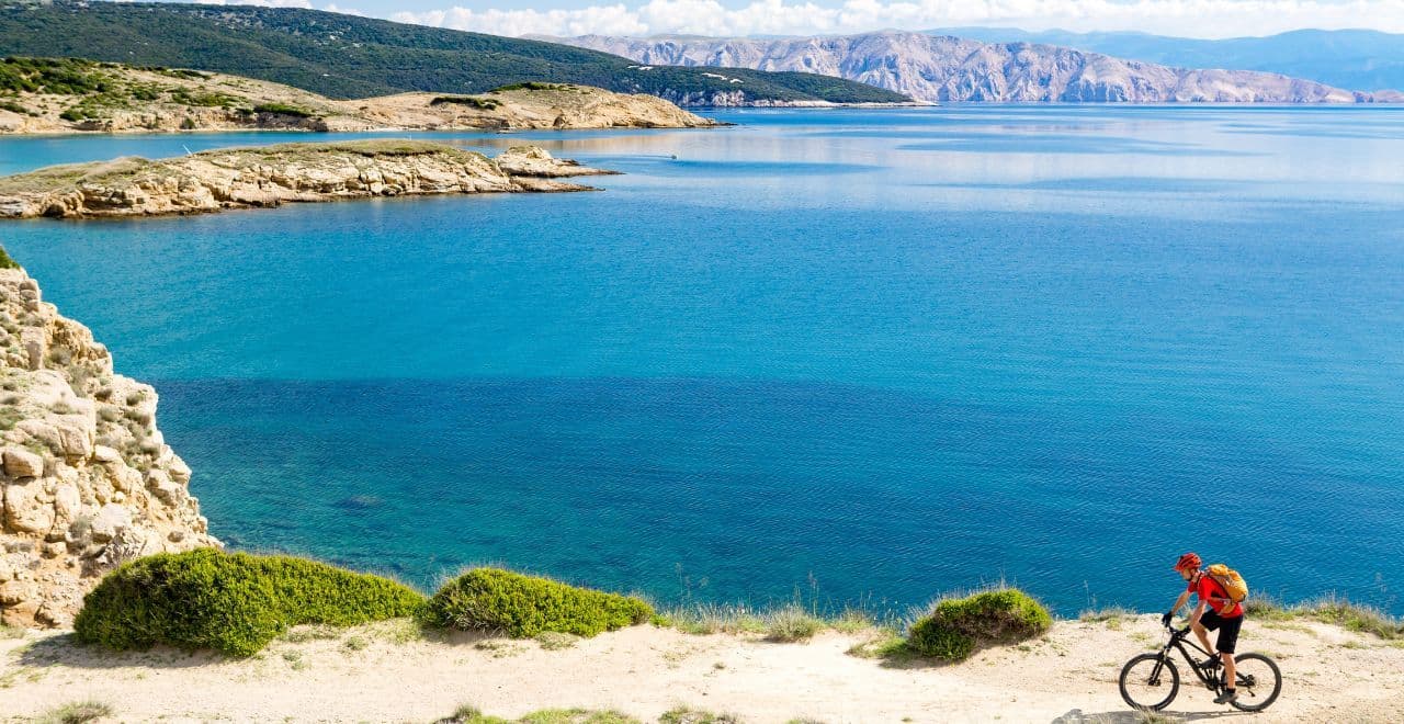 Solo cyclist riding on a rugged coastal path overlooking crystal-clear blue waters, a perfect blend of challenge and natural beauty.