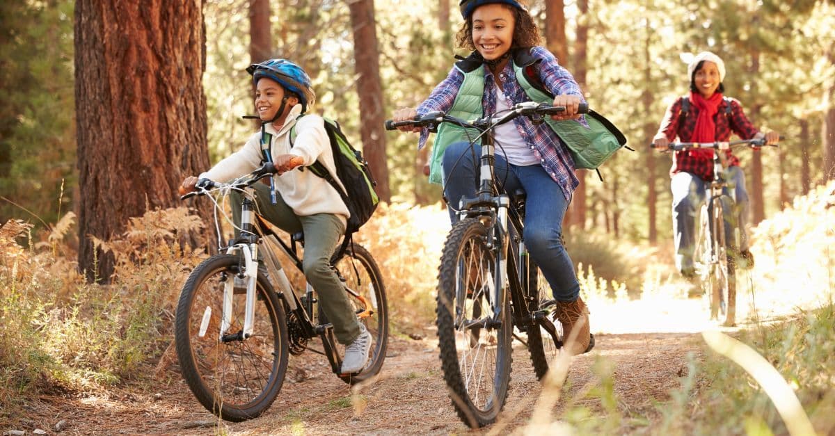 Three young cyclists smiling and enjoying a family bike ride through a sun-dappled forest.