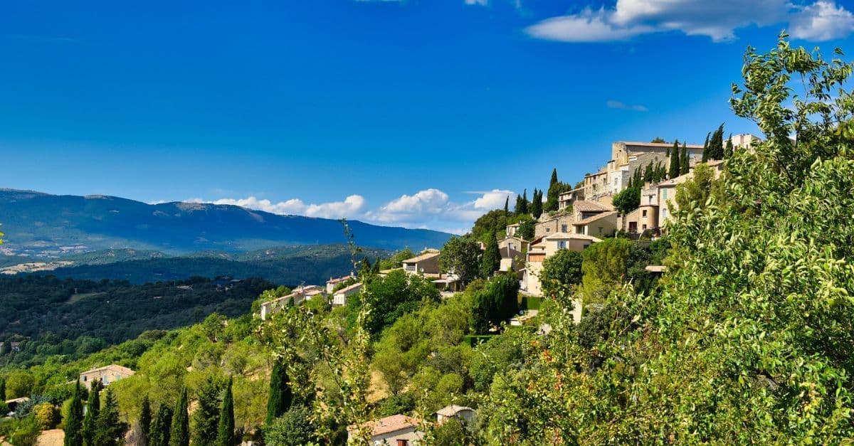 Hillside view of a quaint village in Provence, France, with lush greenery and mountain backdrop