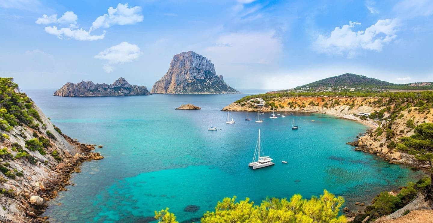 Scenic bay in Ibiza with turquoise waters, sailboats anchored, and rocky islands in the background under a bright blue sky.