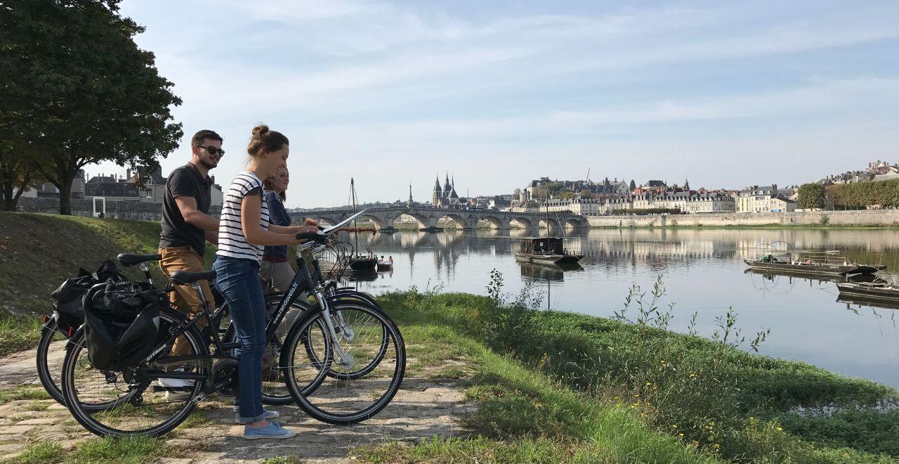 Group of cyclists with a map by a river, with a historic city and bridge visible.