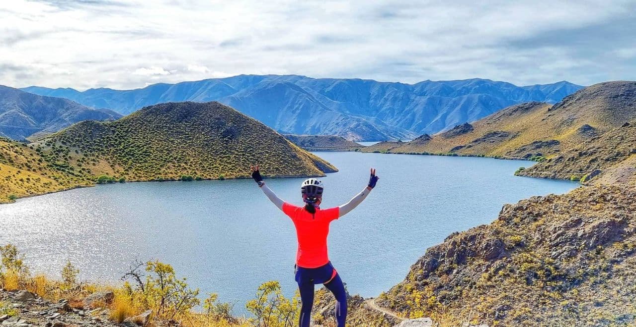 Cyclist in red jersey celebrating at Lake Benmore with mountains in the background.