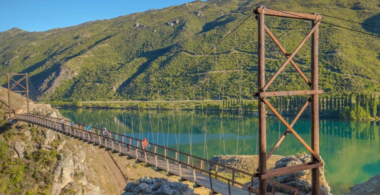 Cyclists crossing a suspension bridge over a scenic lake on the Alps 2 Ocean Cycle Trail.