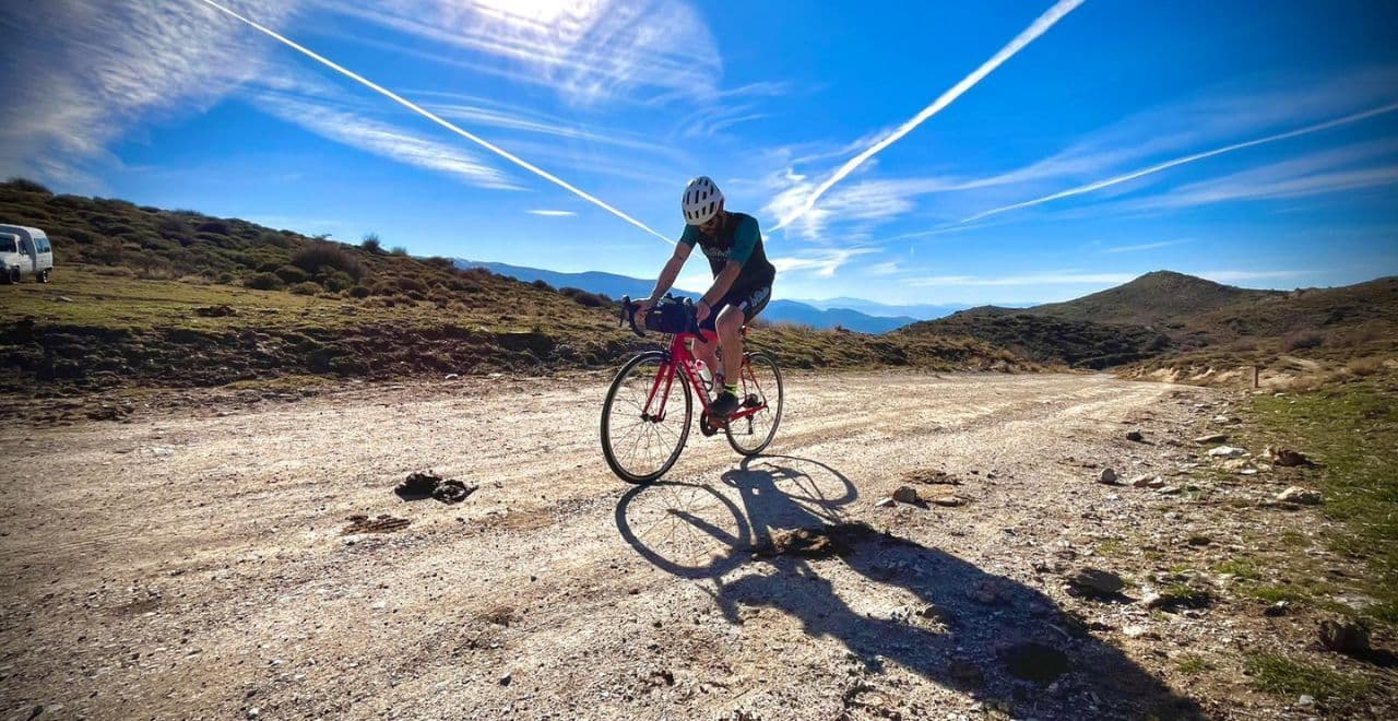 Cyclist on a gravel path in the mountains with bright sunshine and contrail-streaked sky.