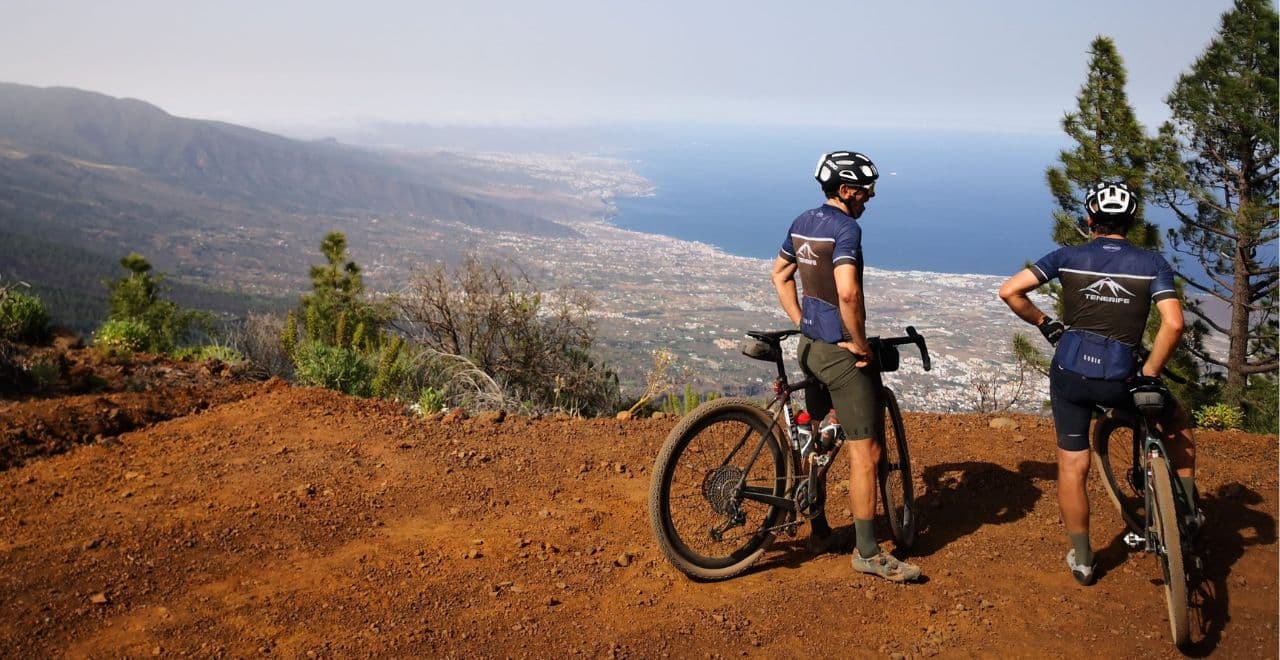 Two cyclists enjoying the scenic view from a high vantage point in Tenerife, with a panoramic view of the coastline and town below.