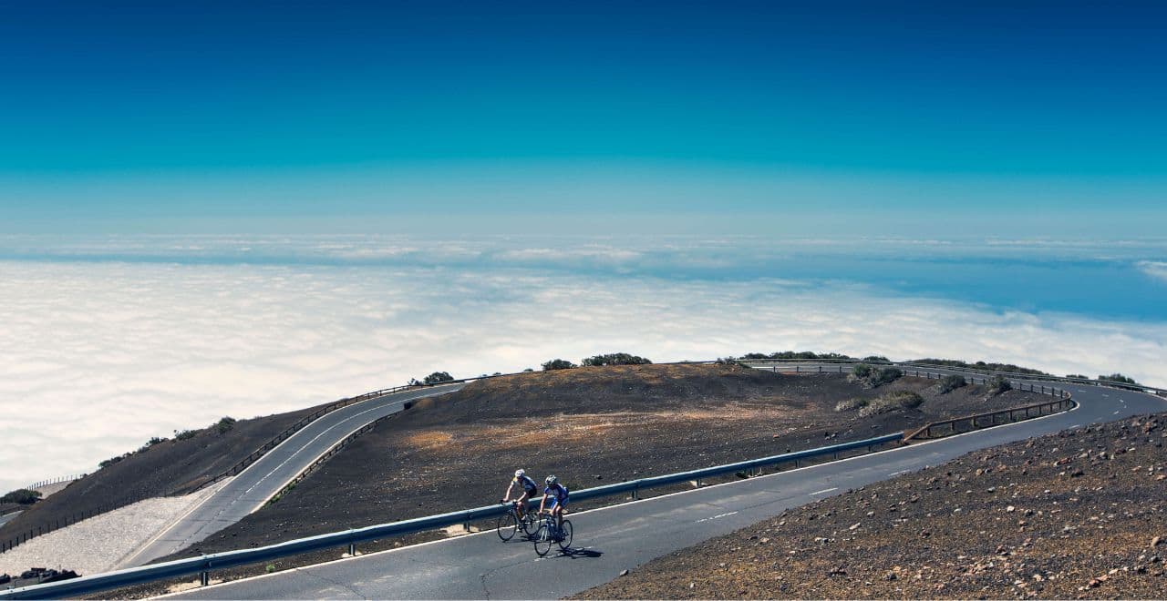 Two cyclists riding on a winding road above the clouds in Tenerife, with a clear blue sky and barren volcanic landscape