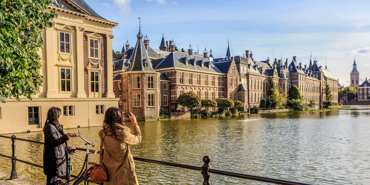 2 ladies standing on a bridge with 1 photographing buildings next to the river