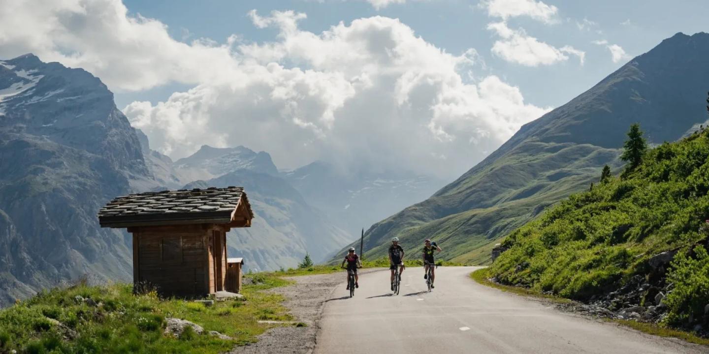 Three cyclists ride along a winding road through the lush green valleys of Col de l'Iseran in the French Alps, with snow-capped mountains in the background under a dramatic sky.