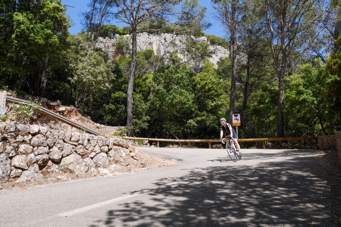 A cyclist navigating a sharp turn on the scenic Coll d'Honor route in Mallorca, surrounded by lush greenery and rocky landscapes.